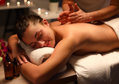 Passive girl lye on massage table covered with white towel and enjoy. Man do back massage with his hand. Candle burn in background.